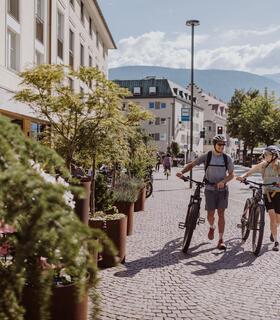 A man and a woman are pushing their bicycles through Bruneck