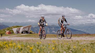 Two cyclists are on the Kronplatz, with the Messner Mountain Museum behind them